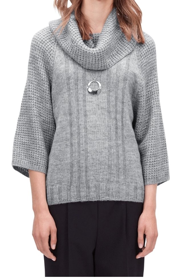 Femme portant un pull en maille gris avec un large col châle, des manches trois-quarts, un pendentif circulaire en argent et un pantalon noir.