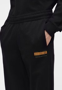 Άτομο που φοράει μαύρη φόρμα Emporio Armani με ελαστική μέση και χέρια στις πλαϊνές τσέπες, πάνω σε ανοιχτό φόντο.