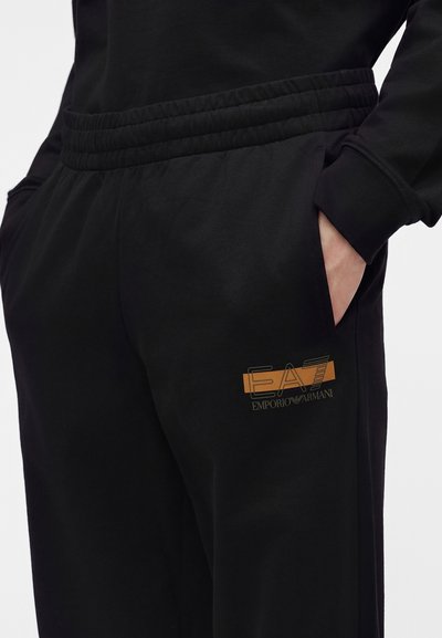 Човек, носещ черни спортни панталони Emporio Armani с еластична талия и ръце в страничните джобове срещу светъл фон.