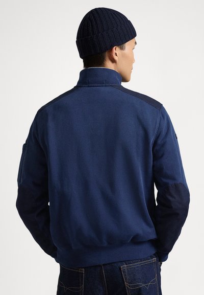 Mornarsko moder flis jopica z visokim ovratnikom, kontrastnimi temnimi rokavi, rebrastimi manšetami in gladko teksturo. Nosljena s temno pleteno kapo.