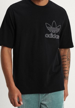 Mężczyzna w czarnej koszulce Adidas z białym logo na piersi, jasno-niebieskich spodniach i widocznym wytatuowanym prawym przedramieniu.