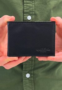 Portefeuille en cuir noir avec une finition texturée, affichant un logo gaufré, tenu entre des mains sur un fond de chemise verte.