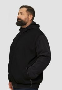 Schwarze Jacke mit strukturiertem Körper und glatten Ärmeln, ausgestattet mit einer Kapuze, Reißverschlusstaschen und verstellbaren Kordeln, die ein kontrastierendes Design schaffen.