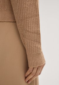 Primo piano di una mano appoggiata di lato, che indossa una manica di maglione a collo alto color cammello e una gonna o pantaloni in tessuto lucido abbinati.