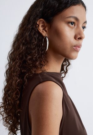 Braunes ärmelloses Top mit glatter Textur. Das lockige Haar des Models fällt über ihre Schulter und wird von großen silbernen Creolen ergänzt.