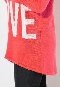 Strickpullover in lebhaftem Koralle mit fettem weißen "LOVE"-Text. Weiche Textur, leicht verlängertem Saum, kombiniert mit dunklen Jeans.