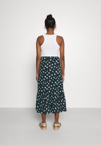 Frau, die mit dem Rücken steht, trägt ein ärmelloses weißes Top, einen schwarzen Midirock mit Blumenmuster in Blau und beige Sandalen.
