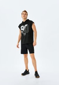 Zwarte mouwloze hoodie met een grote witte "90" graphic en tekst, gecombineerd met zwarte shorts en zwarte sportschoenen. Glad stoffen textuur.