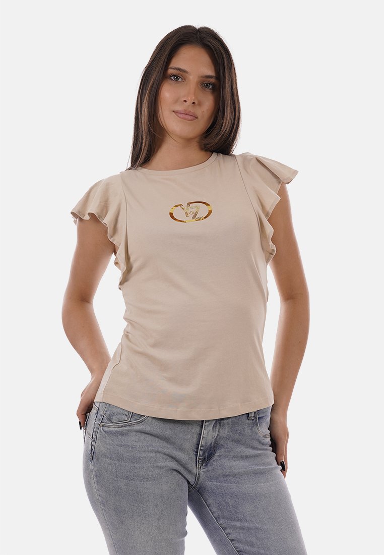 Femme portant un haut beige à manches à volants avec un logo doré sur la poitrine et un jean gris clair, debout les mains dans les poches.