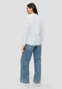 Camisa blanca de manga larga con una textura suave, que presenta un diseño entallado y costuras en la parte trasera, combinada con pantalones vaqueros anchos de mezclilla azul y zapatillas blancas.