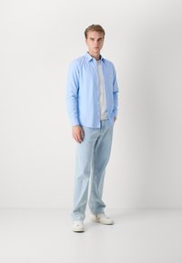 Camicetta azzurro chiaro indossata sopra una maglietta bianca a girocollo, abbinata a jeans azzurri chiari e sneakers bianche. Design semplice, senza motivi.