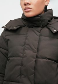 Brūns polsterēts mētelis ar augstu apkakli, kuram ir redzami šuvumi, snap aizdares un gluda, spīdīga tekstūra.