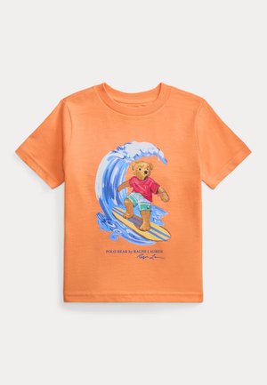 T-shirt orange à manches courtes avec un motif représentant un ours en peluche surfant sur une vague bleue, portant un t-shirt rouge et un short vert.