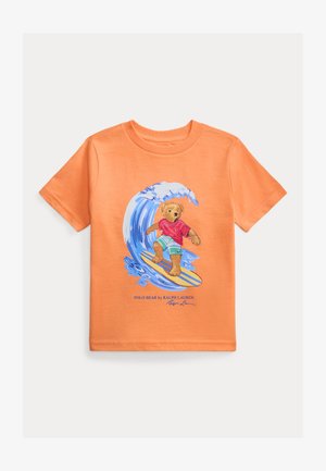 Orange short-sleeve T-shirt with graphic of teddy bear surfing a blue wave, wearing red shirt and green shorts.