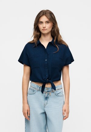 Młoda kobieta z długimi brązowymi włosami, ubrana w granatową koszulę z krótkim rękawem, zapinaną na guziki i zawiązaną z przodu oraz jasnoniebieskie, wysoko taliowane jeansy z łańcuchowym paskiem.