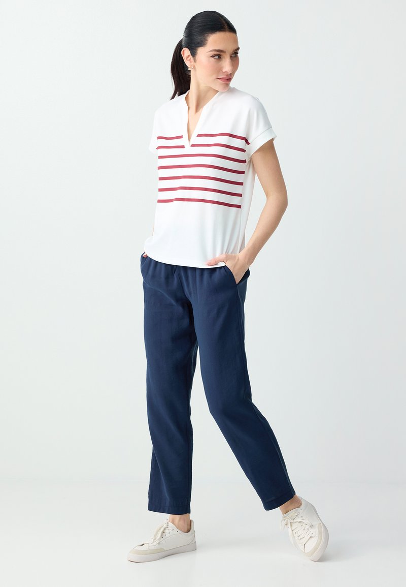 Femme debout portant un haut blanc à manches courtes avec des rayures rouges, un pantalon bleu marine et des baskets blanches, les mains dans les poches, regardant vers la droite.