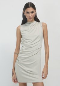 Femme aux cheveux bruns portant une robe grise claire à volants sans manches, se tenant devant un fond blanc uni avec une expression neutre.