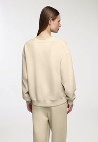 Felpa beige oversize con scollo rotondo e spalle scese, caratterizzata da una texture liscia e un orlo a coste, abbinata a pantaloni coordinati.