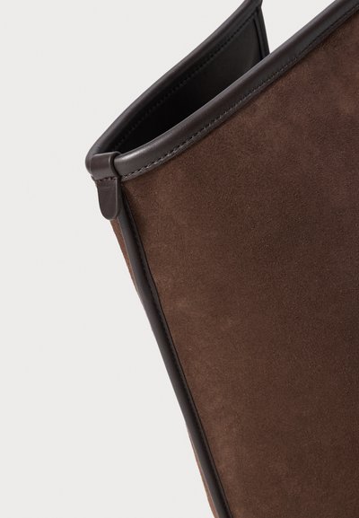 Detailný záber na hnedú semišovú časť topánky s tmavou koženou obrubou a vyšívaním na bielom pozadí.