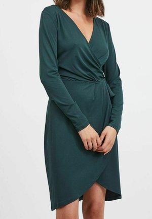 Smaragdgroene wikkeljurk met lange mouwen, V-hals, geplooide taille en een asymmetrische zoom tot op de knie. Gemaakt van zachte, rekbare stof.