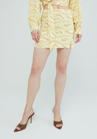 Geel gemusterde minirok met een strik in de taille, met een hoge split. Gecombineerd met bruine open teen hakken.