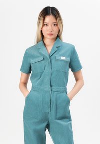 Teal kordfløyelsdress med korte ermer, to brystlommer og en tilpasset midje. Har subtile vertikale striper og front knappelukking.