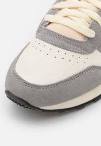 Sko med en överdel i beige läder, grå mockadetaljer och perforerade inslag. Har krämfärgade snörningar och en svart gummisula.