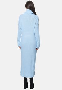 Langes, hellblaues ribbeltes Strickkleid mit hohem Kragen und langen Ärmeln, das seitliche Schlitze und eine lockere Passform aufweist. Tragen mit schwarzen Plateaustiefeln.