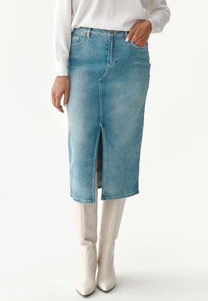 Femme portant une jupe midi en denim bleu clair avec une fente à l'avant, un top blanc côtelé et des bottes blanches pointues montant aux genoux.