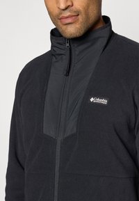 Black fleece jacket with a high collar, front zipper, and a smooth fabric panel on the chest. Features logo on the left side.
