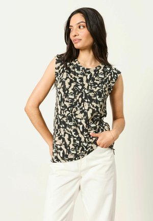 Femme aux longs cheveux foncés portant un haut sans manches à motifs noirs et beige et un pantalon blanc, posant avec une main dans la poche.