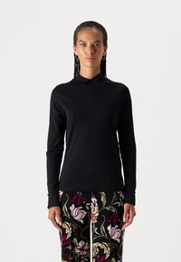 Schwarzes langärmliges Oberteil mit Stehkragen, glatter Textur und taillierter Passform, kombiniert mit gemusterten, bunten Blumenhosen.