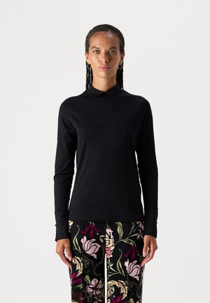 Schwarzes langärmliges Oberteil mit Stehkragen, glatter Textur und taillierter Passform, kombiniert mit gemusterten, bunten Blumenhosen.