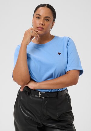 Femme aux cheveux foncés portant un t-shirt bleu avec un petit motif de cœur et un pantalon en cuir noir, debout la main sur le menton.
