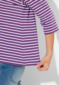 Lila gestreiftes Hemd mit weißen horizontalen Linien, mit dreiviertel Ärmeln und geradem Saum. Die Hand des Models hält den unteren Rand.