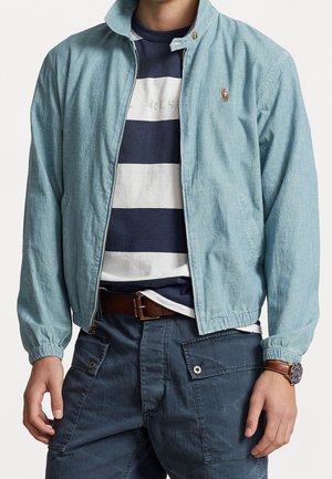 Mężczyzna w jasnoniebieskiej kurtce zapinanej na zamek, nałożonej na granatowo-białą pasiastą koszulkę, ciemnych spodniach typu cargo, brązowym skórzanym pasku i zegarku na nadgarstku.