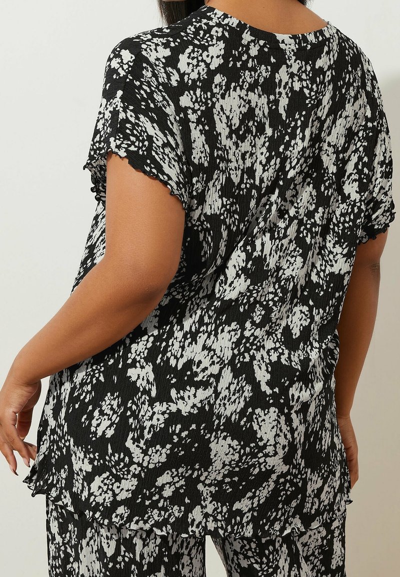 Femme portant un chemisier court à fleurs noir et blanc et un pantalon assorti, vue de dos sur un fond neutre.