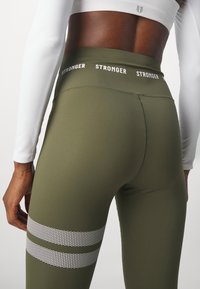 Leggings vert olive avec une large ceinture comportant le mot "STRONGER" en blanc. Accents texturés et brillants dans un motif de rayures horizontales.