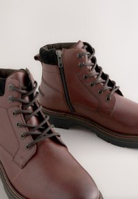 Braune Leder-Stiefeletten mit strukturierter Oberfläche, schwarzem Stoffkragen und seitlichem Reißverschluss. Verfügen über runde Schnürsenkel und eine robuste Gummisohle.