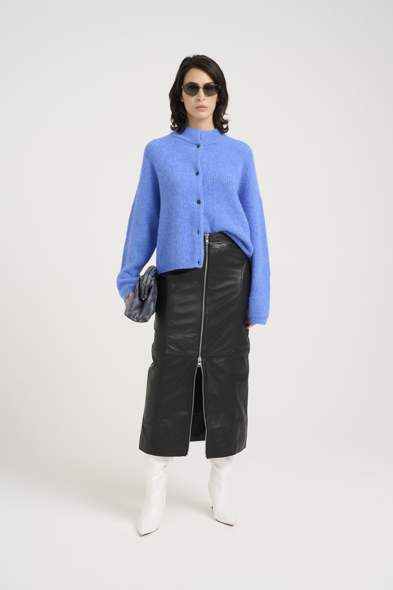 Blaue gestrickte Strickjacke mit Knöpfen, schwarzer Lederrock mit Frontreißverschluss, weiße spitze Stiefel, graue strukturierte Clutch und Sonnenbrille.
