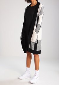 Cardigan à motifs noir et blanc avec une longue ouverture frontale, manches larges, deux poches, porté sur une robe noire, associé à des baskets blanches.