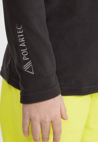 Veste en polaire noire avec le logo Polartec sur la manche, présentant une texture lisse et un design à col rond. Pantalons jaunes vifs visibles en arrière-plan.