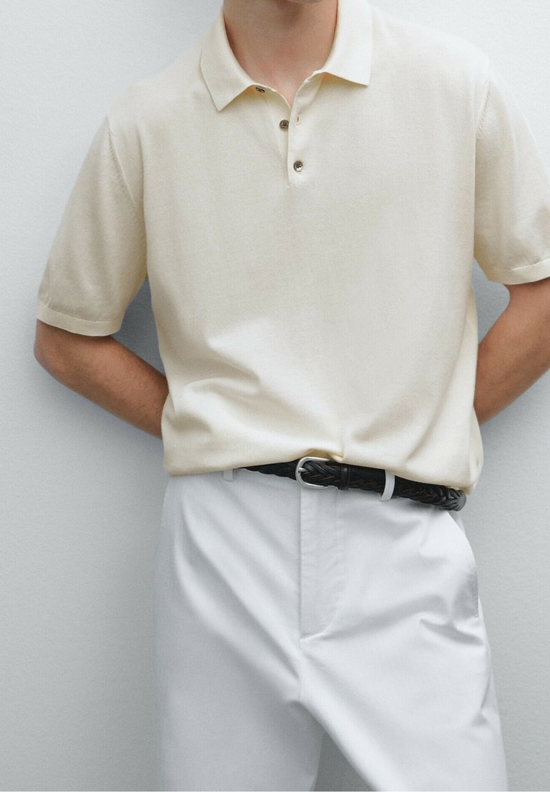 Polo beige à trois boutons, manches courtes et texture lisse, associé à un pantalon blanc et une ceinture noire tressée.