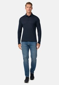 Hombre vistiendo una camiseta polo de manga larga azul marino, jeans azules y zapatillas negras, de pie contra un fondo liso gris claro.