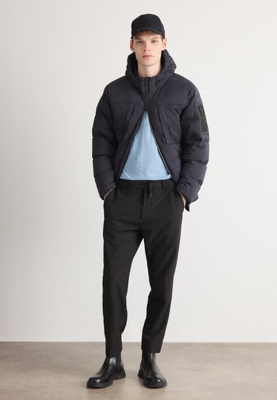 Veste noire rembourrée avec capuche, t-shirt bleu et pantalon noir. Complété par des bottines noires et une casquette noire. Design simple et moderne.