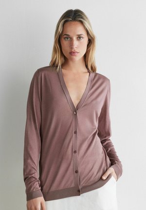 Kvinde med blondt hår iført en lilla knaptrøje og hvid nederdel, stående mod en ensfarvet lys grå væg.