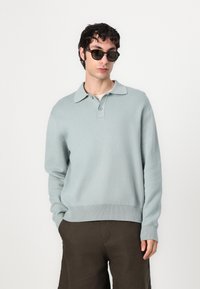 Light blue knit pullover with a collar, button detail, and ribbed hem. Paired with dark shorts. Model wears dark sunglasses.
