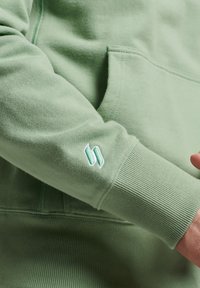 Grön hoodie, gjord av mjukt tyg, med en känguruficka och ribbade muddar. Vänster ärm har en broderad logotyp i en ljusare grön nyans.