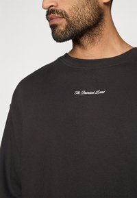 Melna džemperis ar apaļo kakla izgriezumu, mīkstu tekstūru un izšūtu tekstu "Solītā zeme" uz krūtīm.