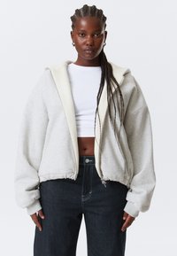 Kvinde med lange fletninger iført en oversized lysegrå hættetrøje med lynlås over en hvid crop top og mørke højtaljede jeans, stående mod en hvid baggrund.